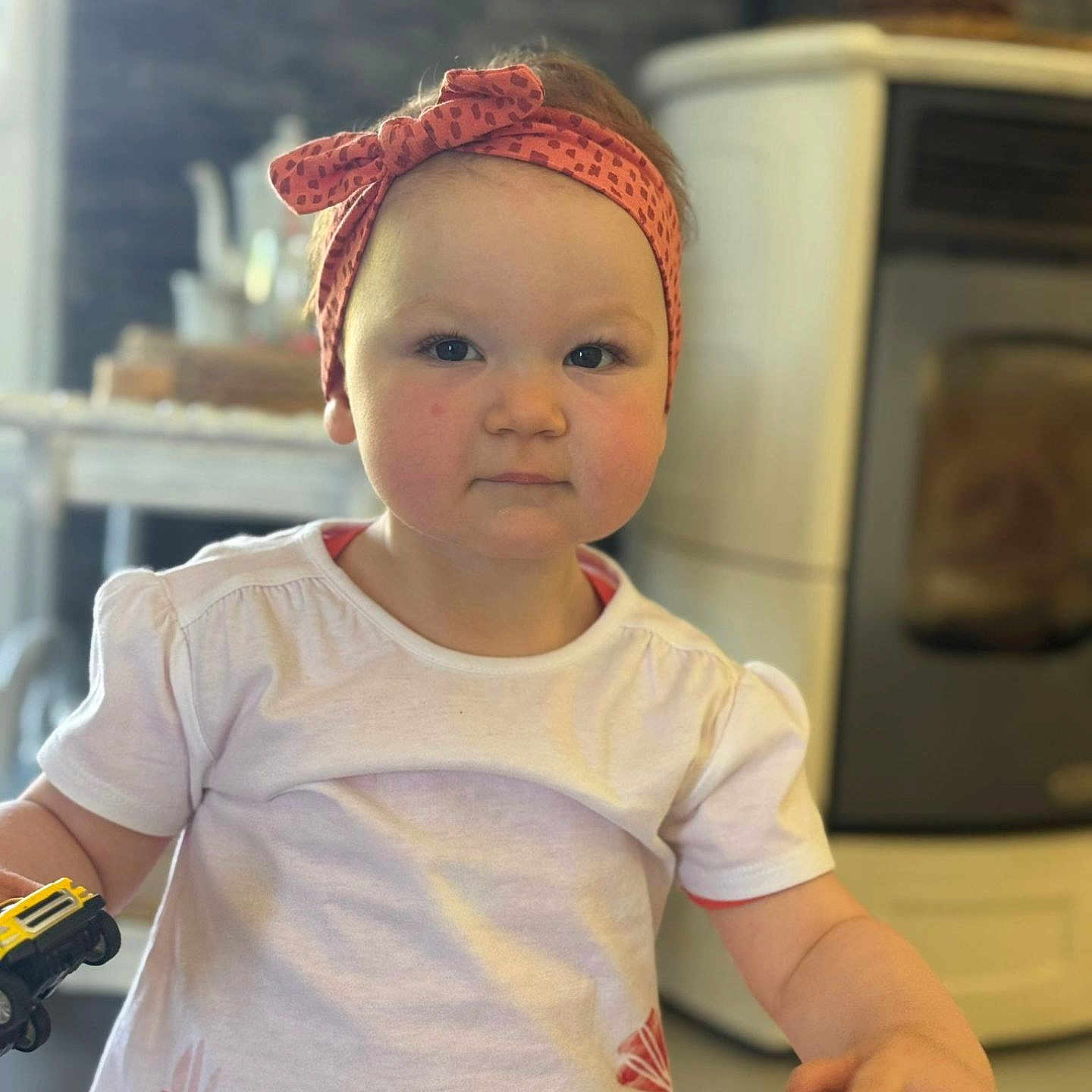 Rose a rejoint le concours — aidez-le/la à gagner de superbes lots ! blurred_background, child, cute, domestic, dress, face, floral_pattern, furniture, headband, holding, indoor, person, pink, playing, short_sleeve, stove, table, toddler, toy_car, young_child
