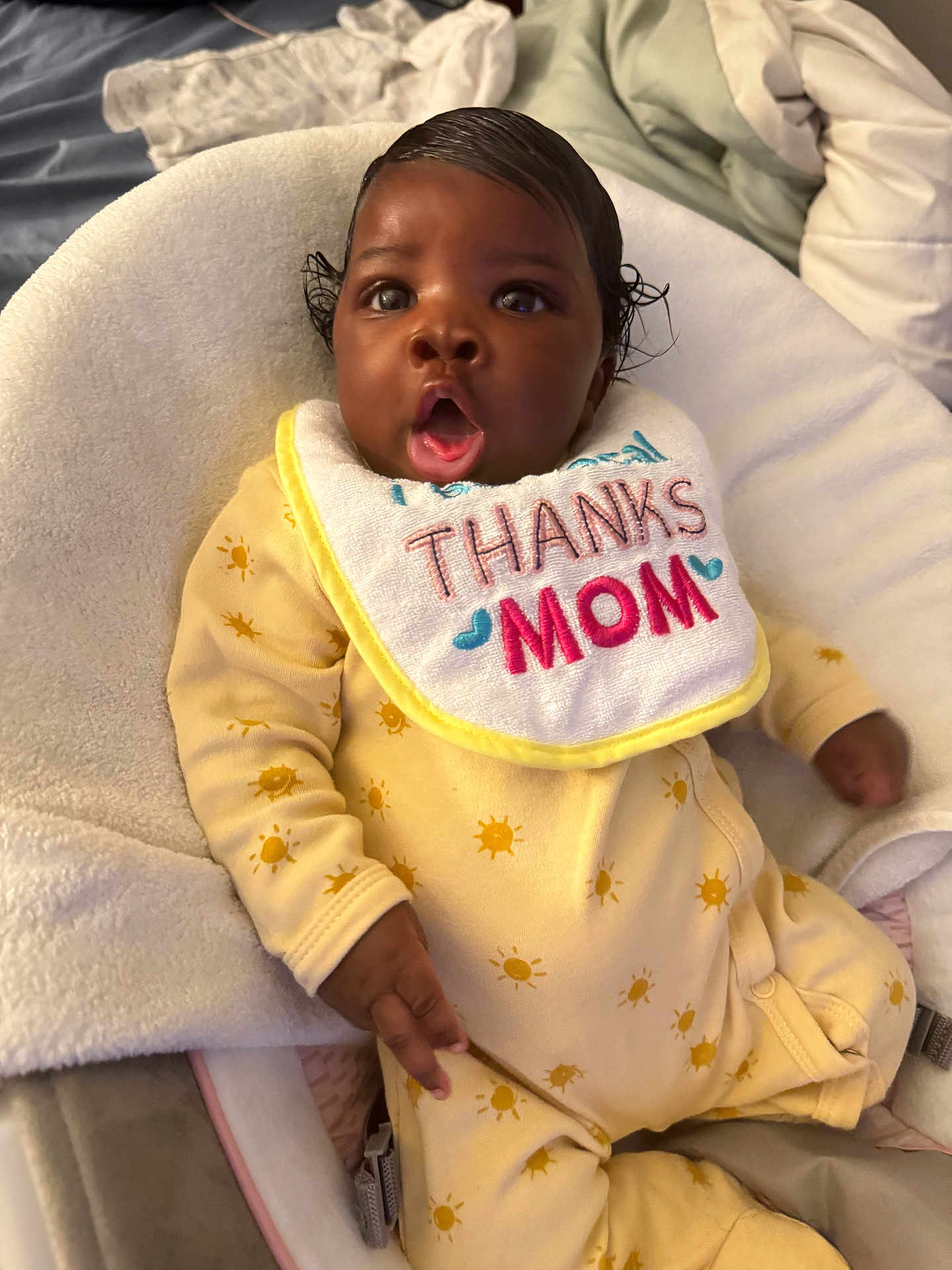 Maelise is registered to the contest to win money with this photo: baby, child, infant, onesie, yellow_clothing, bib, thanks_mom, baby_seat, blanket, expression, surprised, indoor, soft_texture, cushion, human, person, cute, portrait, skin, hand