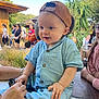 baby, child, cap, outdoor, people, hand, blue_clothing, smiling, grass, trees, wood, table, glasses, person, vape, sitting, daylight, nature, casual, happy