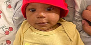 Kai Green joined the competition — help win amazing prizes! baby, child, red_beanie, yellow_shirt, text_on_clothing, blanket, pattern, cute, smiling, lying_down, indoor, person, infant, young_child, headwear, apparel, face, hand, soft_lighting, cozy