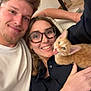 couple, smiling, indoor, selfie, cat, kitten, orange_cat, glasses, black_shirt, white_shirt, happy, playful, tongue_out, close_up, human_face, pet, casual, relaxed, arm, home