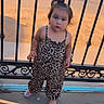 Naylan is registered to the contest to win money with this photo: child, toddler, leopard_print, jumpsuit, standing, fence, metal_fence, outdoor, concrete, sidewalk, sunlight, shadow, street, bracelet, footwear, hair_tie, curious, young_child, daylight, person
