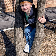 Blake is registered to the contest to win money with this photo: baseball_cap, boy, brick_wall, cap_backwards, casual_clothing, child, hands, jeans, nature, outdoor, peeking, playground, portrait, red_hair, shadow, smile, sneakers, sunlight, tree, tree_trunk