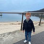 child, boy, red_hair, smiling, standing, jacket, jeans, sneakers, waterfront, pavement, railing, overcast, outdoor, nature, sky, water, beach, fence, person, casual_clothing