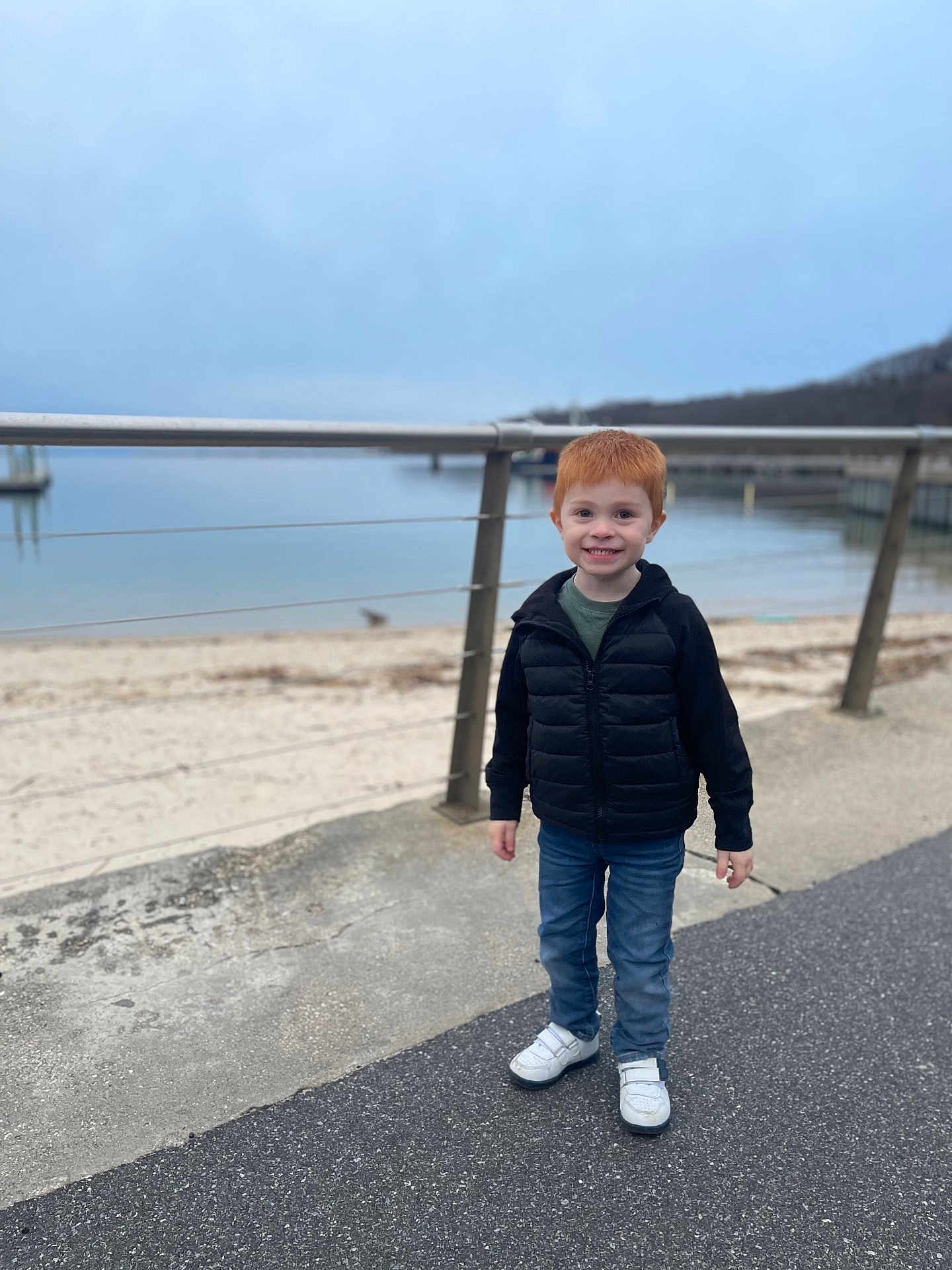 Blake joined the competition — help win amazing prizes! child, boy, red_hair, smiling, standing, jacket, jeans, sneakers, waterfront, pavement, railing, overcast, outdoor, nature, sky, water, beach, fence, person, casual_clothing