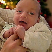 Ezio participe au concours pour gagner de l'argent avec cette photo : baby, child, smile, happy, face, hand, finger, clothing, sweater, indoor, person, infant, expression, closeup, portrait, holding, warm_lighting, background_blur, christmas, decorations