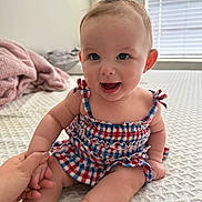 Aliviah is registered to the contest to win money with this photo: baby, child, smiling, sitting, dress, headband, flower, hand_holding, indoor, bed, blanket, window, light, happy, cute, skin, face, foot, person, infant
