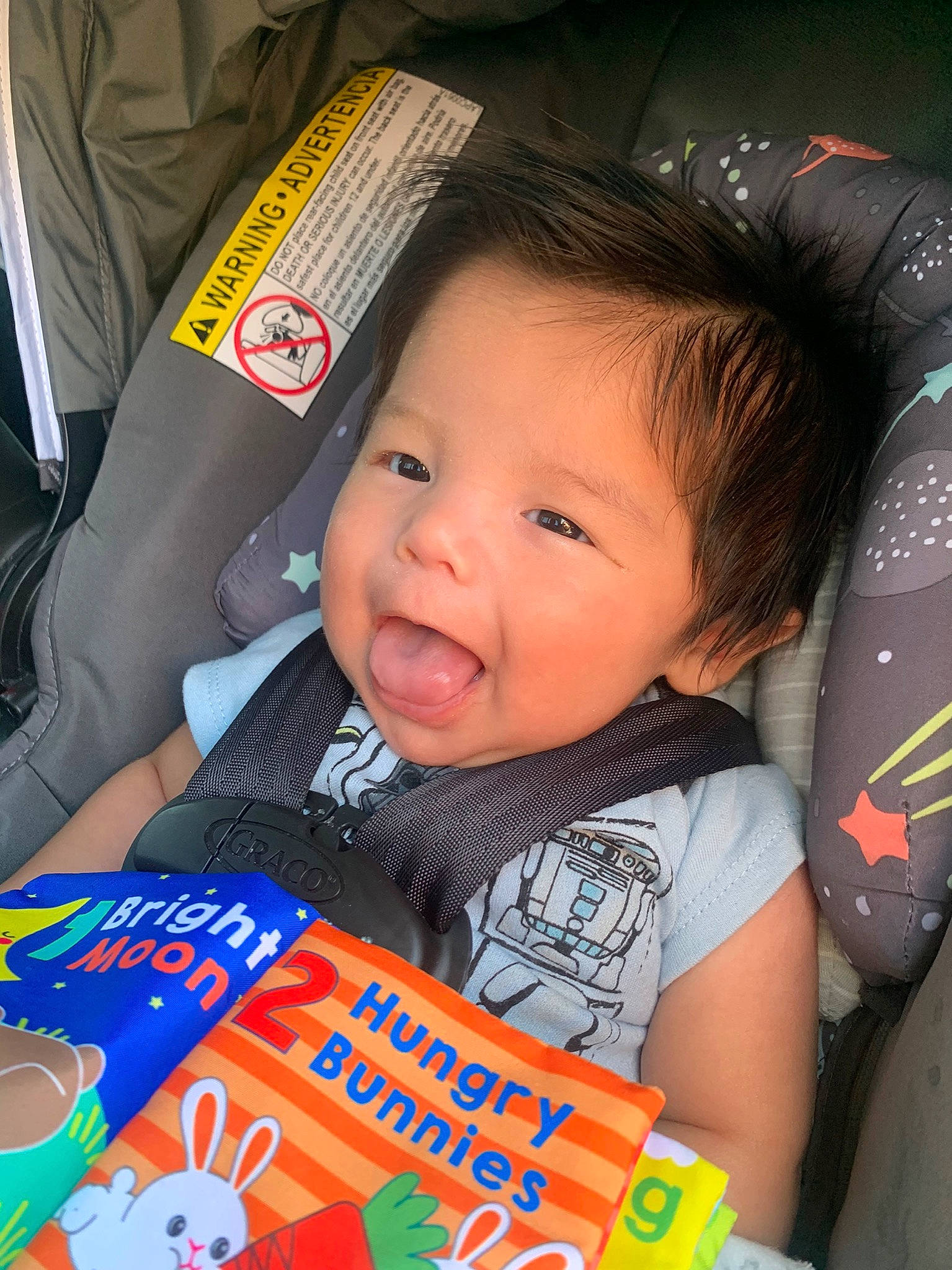 Joseph is registered to the contest to win money with this photo: baby, baby_products, baby_toddler_clothing, book, car_seat, cheek, child, comfort, facial_expression, fun, hairstyle, happy, iris, person, product, publication, reading, sitting, smile, t_shirt