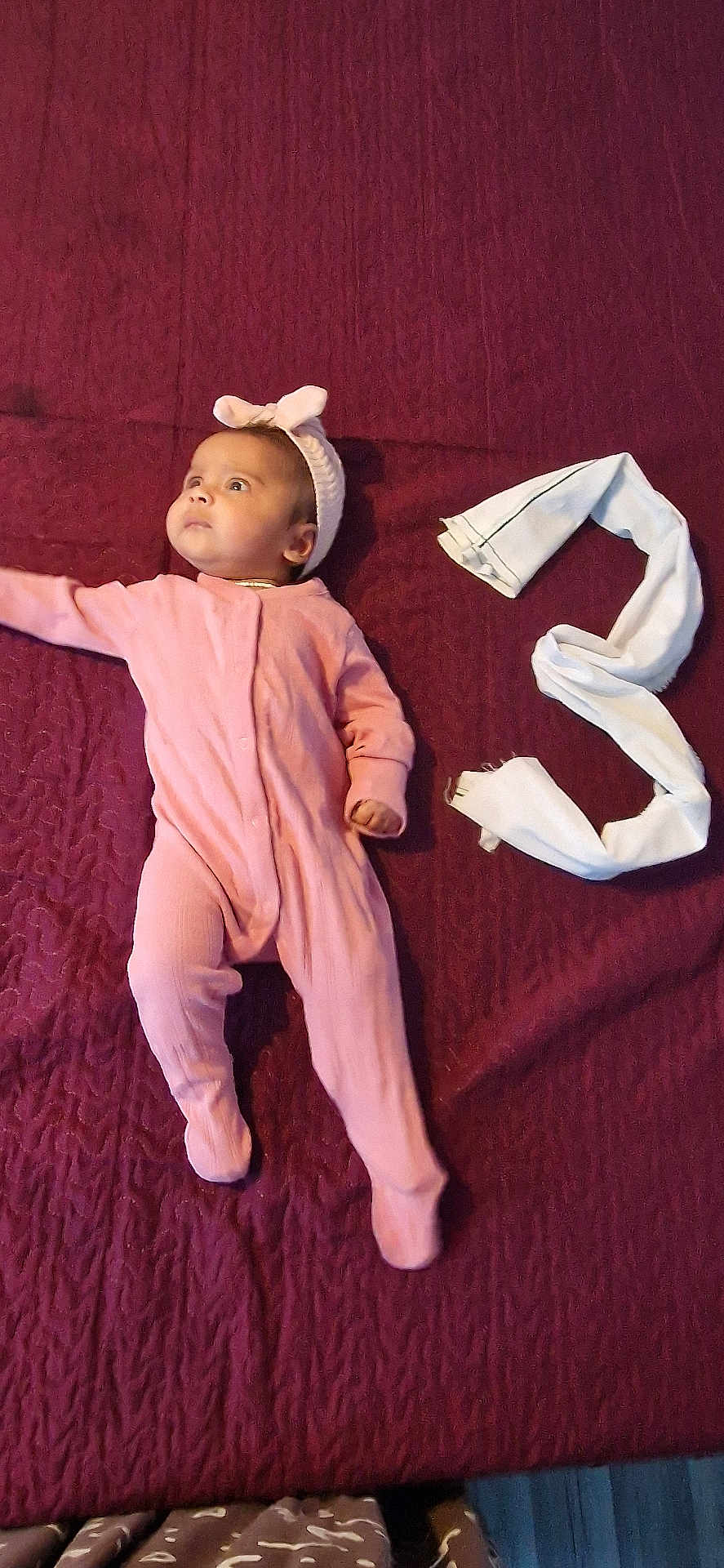 Duwaraka is registered to the contest to win money with this photo: baby, pink_onesie, headband, maroon_blanket, white_cloth, number_three, infant, lying_down, cute, portrait, child, soft_texture, indoors, looking_away, small_hand, footed_pajamas, blanket, fabric, resting, young_child