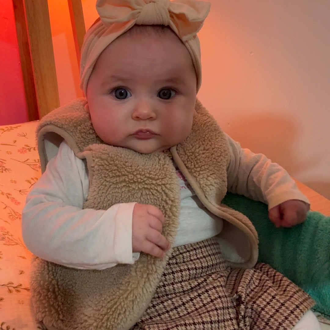 Ambre a rejoint le concours — aidez-le/la à gagner de superbes lots ! baby, child, headband, bow, vest, sitting, bed, lamp, warm_light, curious, plaid_skirt, long_sleeve_shirt, indoors, cozy, furniture, blanket, person, portrait, cute, toddler