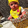 baby, child, yellow_hat, yellow_bib, basket, carrot_pattern, indoor, face, curious, cute, clothing, infant, person, soft_texture, fabric, plush, seated, closeup, expression, headwear