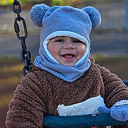 Rayaan is registered to the contest to win money with this photo: child, toddler, smiling, swing, outdoor, hat, mittens, knitwear, blue, brown, playground, happy, face, person, winter_clothing, cute, park, toy, fuzzy, baby