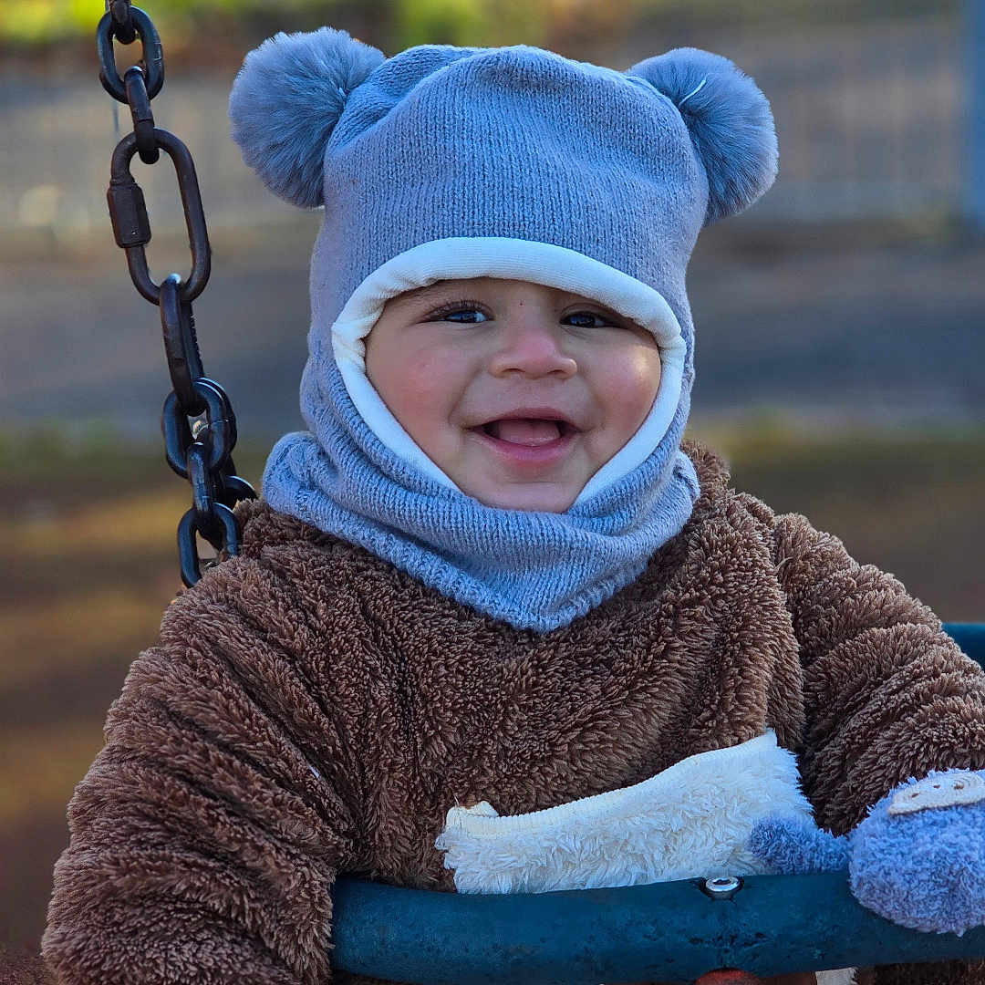 Rayaan is registered to the contest to win money with this photo: baby, blue, brown, child, cute, face, fuzzy, happy, hat, knitwear, mittens, outdoor, park, person, playground, smiling, swing, toddler, toy, winter_clothing