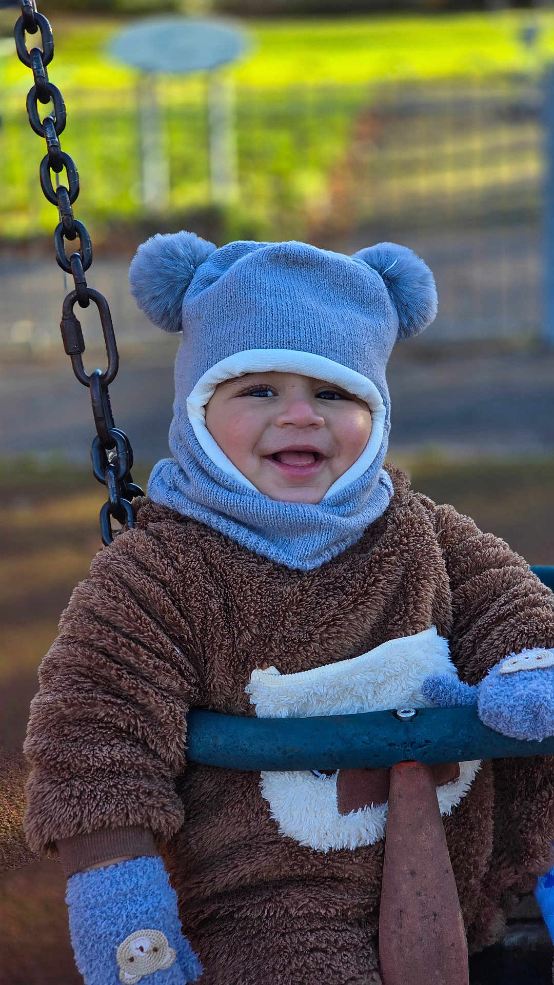 Rayaan is registered to the contest to win money with this photo: child, toddler, smiling, swing, outdoor, hat, mittens, knitwear, blue, brown, playground, happy, face, person, winter_clothing, cute, park, toy, fuzzy, baby