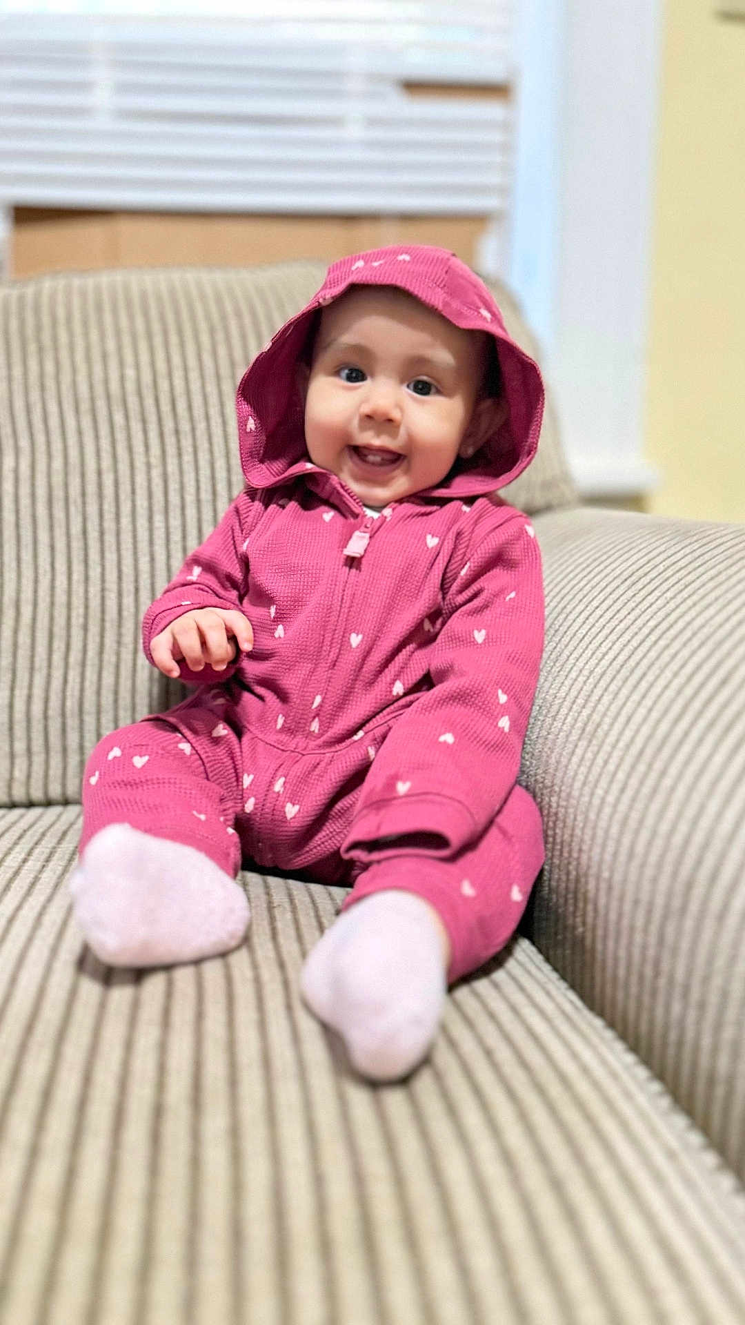 Alayna is registered to the contest to win money with this photo: baby, child, pink_onesie, hooded_clothing, smiling, sitting, couch, striped_fabric, indoor, cute, happy, infant, portrait, cozy, clothing, face, person, feet, socks, home
