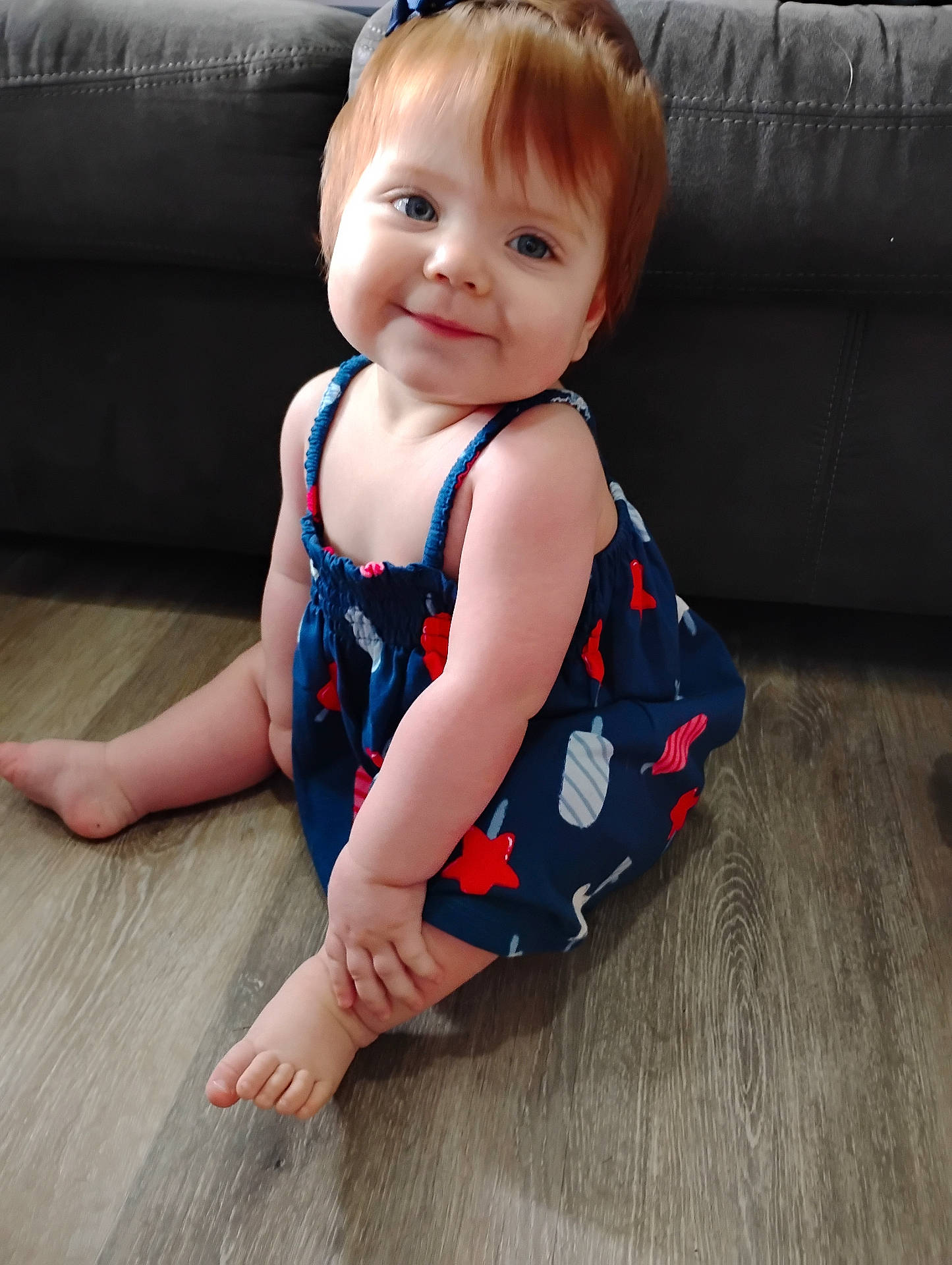 Chloe is registered to the contest to win money with this photo: baby, blue_clothing, child, couch, cute, face, floor, foot, happy, head, home, indoor, person, red_pattern, romper, sitting, skin, smiling, toddler, wooden_floor