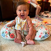 Freya joined the competition — help win amazing prizes! baby, smiling, sitting, indoors, carpet, pillow, floral_pattern, bib, pacifier, child, person, happy, cute, infant, clothing, footwear, toy, living_room, blurred_background, candid