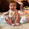baby, smiling, sitting, indoors, carpet, pillow, floral_pattern, bib, pacifier, child, person, happy, cute, infant, clothing, footwear, toy, living_room, blurred_background, candid