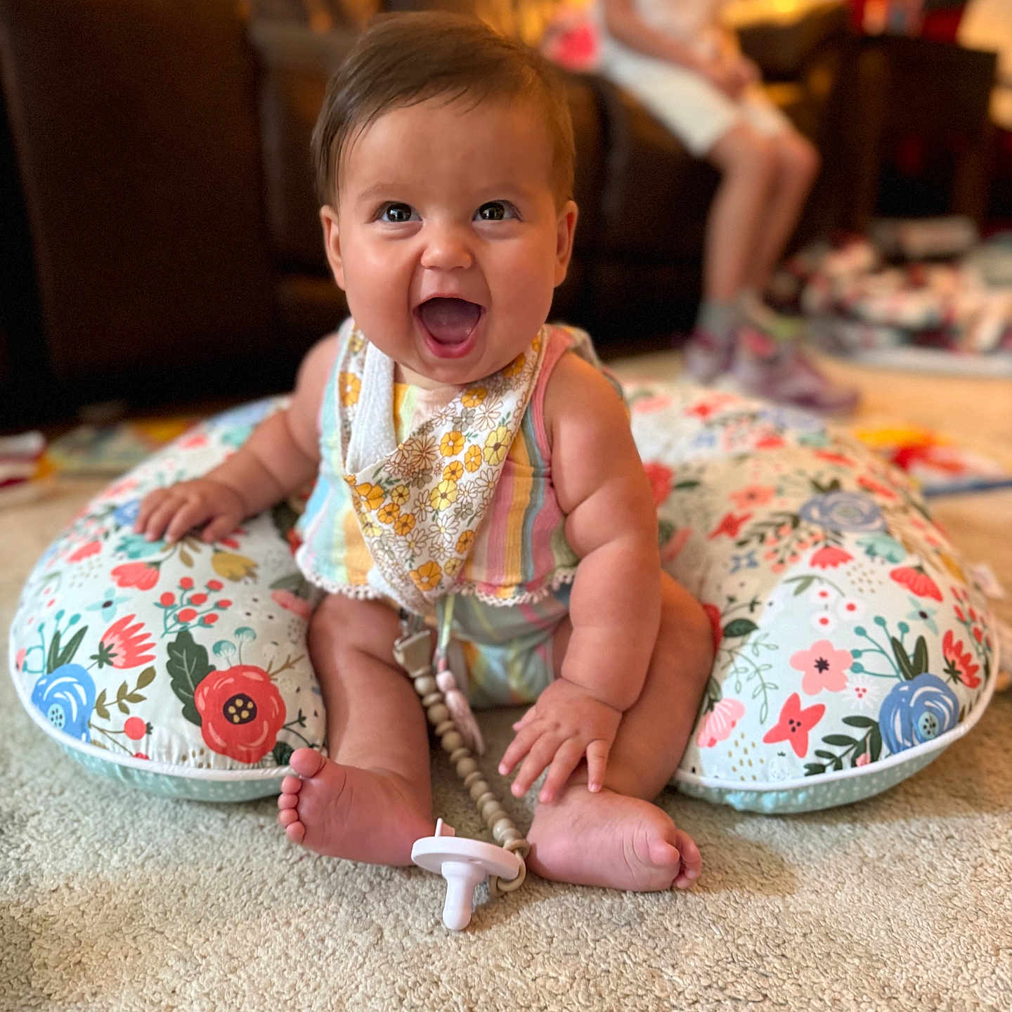 Freya joined the competition — help win amazing prizes! baby, smiling, sitting, indoors, carpet, pillow, floral_pattern, bib, pacifier, child, person, happy, cute, infant, clothing, footwear, toy, living_room, blurred_background, candid