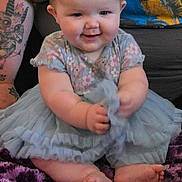 Jaycie is registered to the contest to win money with this photo: baby, child, smiling, blue_dress, tattoo, arm, purple_blanket, sitting, happy, indoor, person, clothing, cute, infant, face, feet, hand, pattern, fabric, toddler
