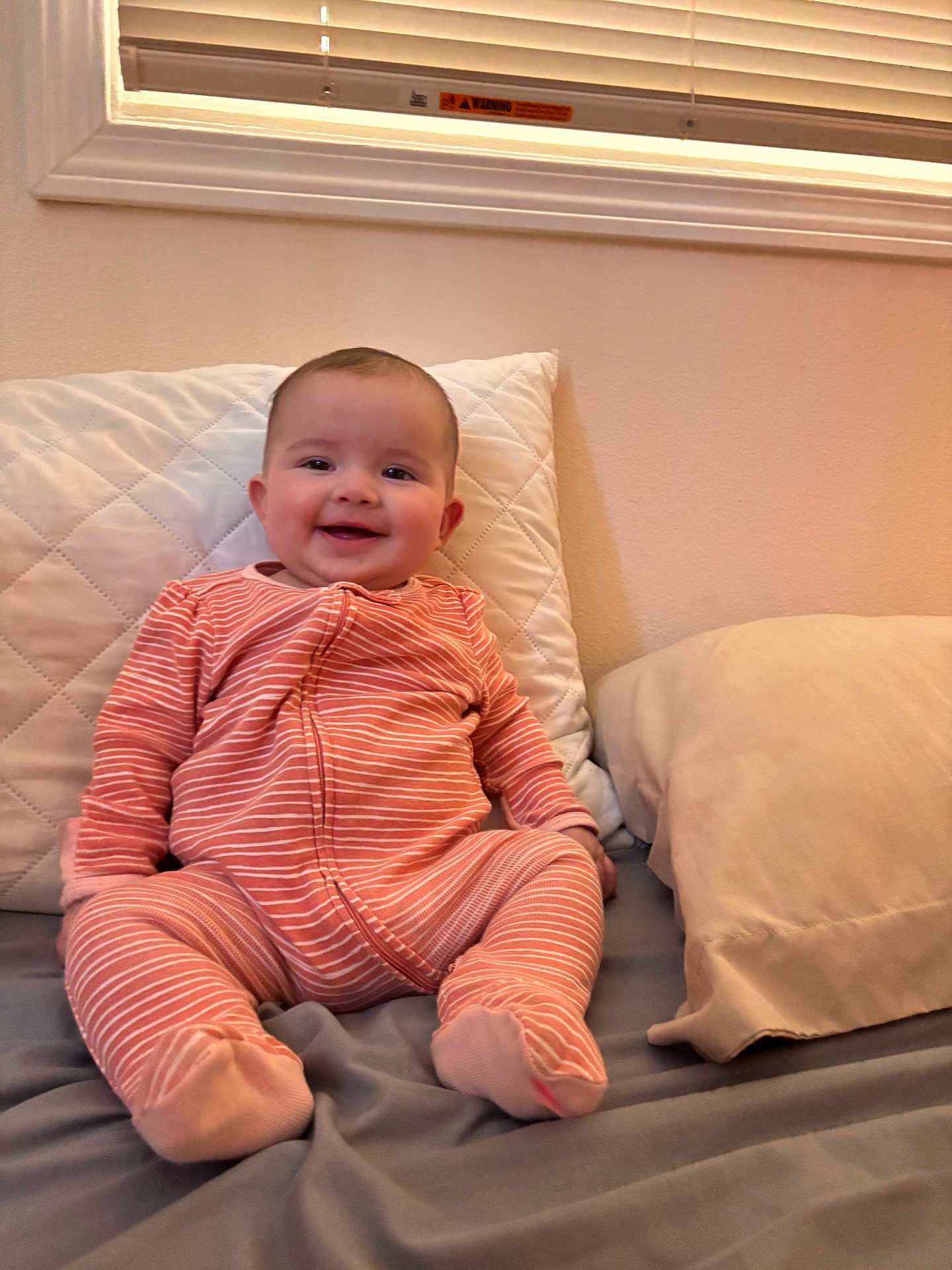 Aixa joined the competition — help win amazing prizes! baby, infant, smiling, pink_onesie, striped_clothing, bed, pillow, indoor, child, happy, cute, person, face, sitting, cozy, home, blanket, soft_lighting, window, curtains