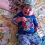 baby, bedspread, blue_clothing, child, colorful, cute, floral_pattern, flower, happy, headband, indoors, infant, joyful, lying_down, owl_design, person, pillow, pink, smiling, soft_lighting