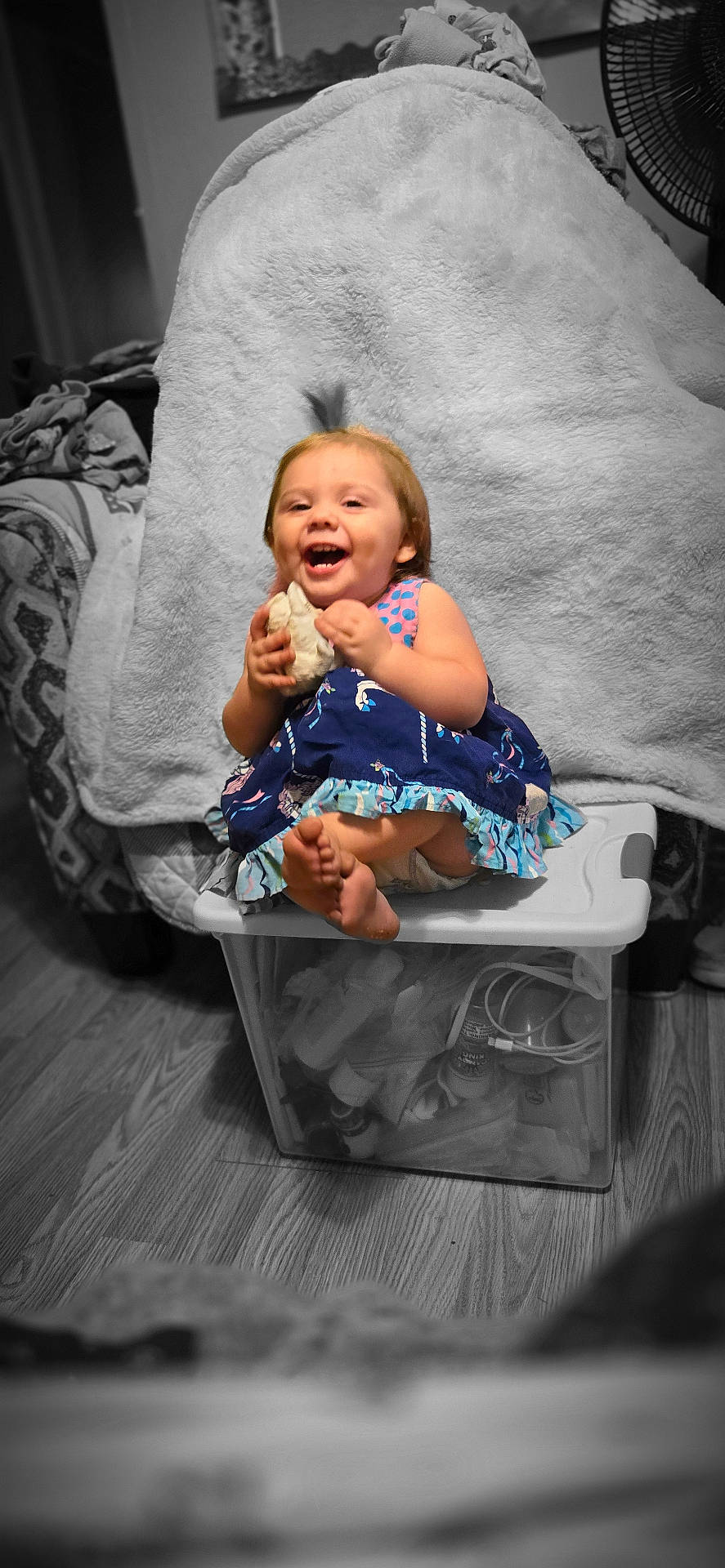 blanket, child, color_pop, cute, dress, fan, floor, foot, happy, holding_object, home, indoor, laughing, person, plastic_container, sitting, smiling, toddler, toy, wooden_floor