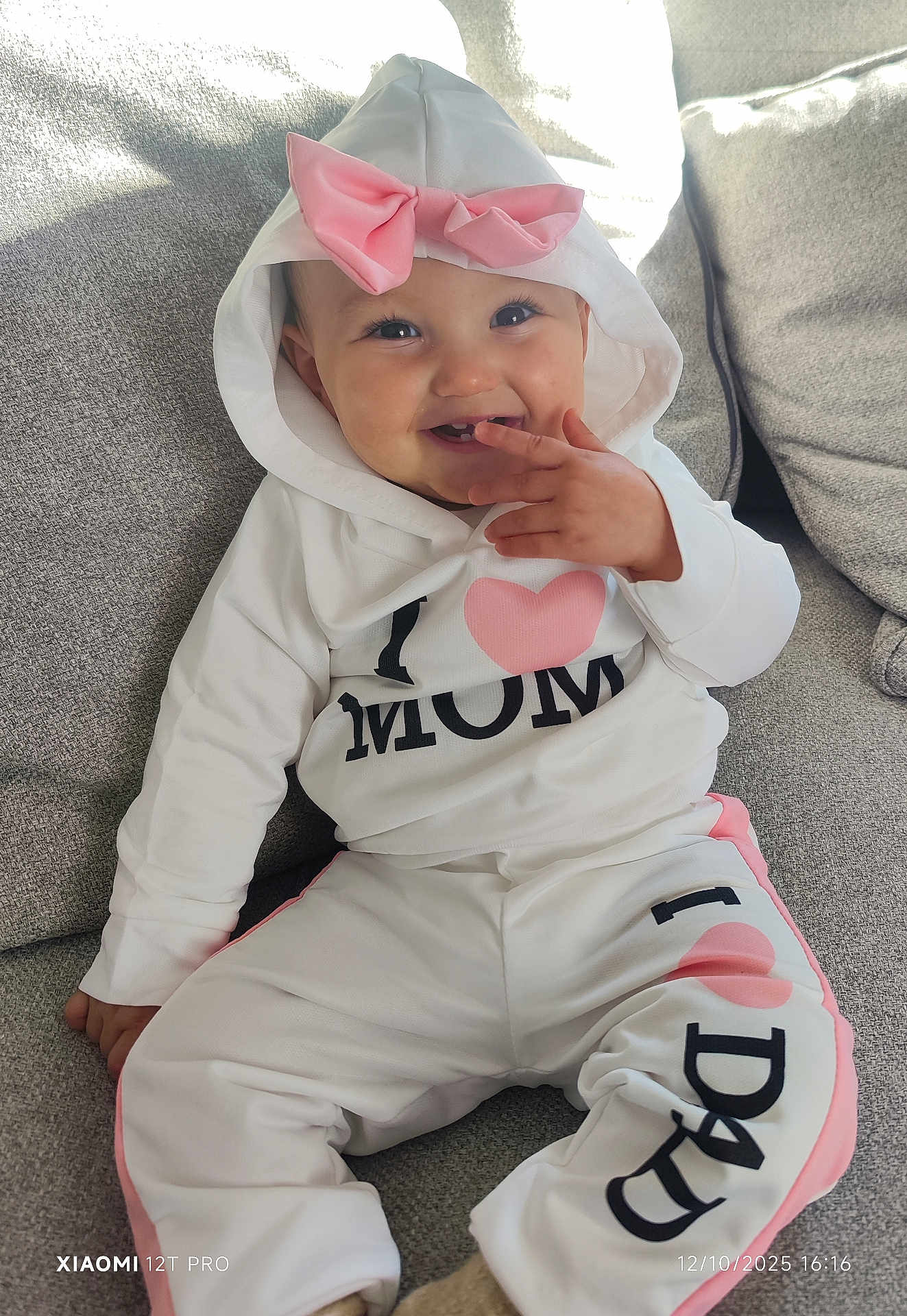 Nina a rejoint le concours — aidez-le/la à gagner de superbes lots ! baby, child, smiling, hoodie, pink_bow, white_clothing, pants, sitting, couch, indoor, happy, cute, infant, face, person, playful, cozy, casual_wear, portrait, toddler