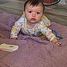 baby, child, crawling, onesie, floral_pattern, purple_quilt, floor, hardwood_floor, indoor, furniture, red_furniture, toy, remote_control, curious, face, person, infant, young_child, expression, cable