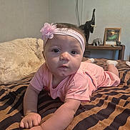 Raelyn is registered to the contest to win money with this photo: baby, child, pink_clothing, headband, flower, blanket, striped_blanket, bed, elephant_figurine, shelf, frame, indoor, person, portrait, cute, lying_down, curious, home, cozy, decor