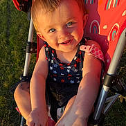 Oakley is registered to the contest to win money with this photo: toddler, child, stroller, outdoor, sunlight, grass, blue_eyes, smiling, happy, playing, summer, rainbow_pattern, clothing, skin, baby, foot, hand, seat, closeup, portrait