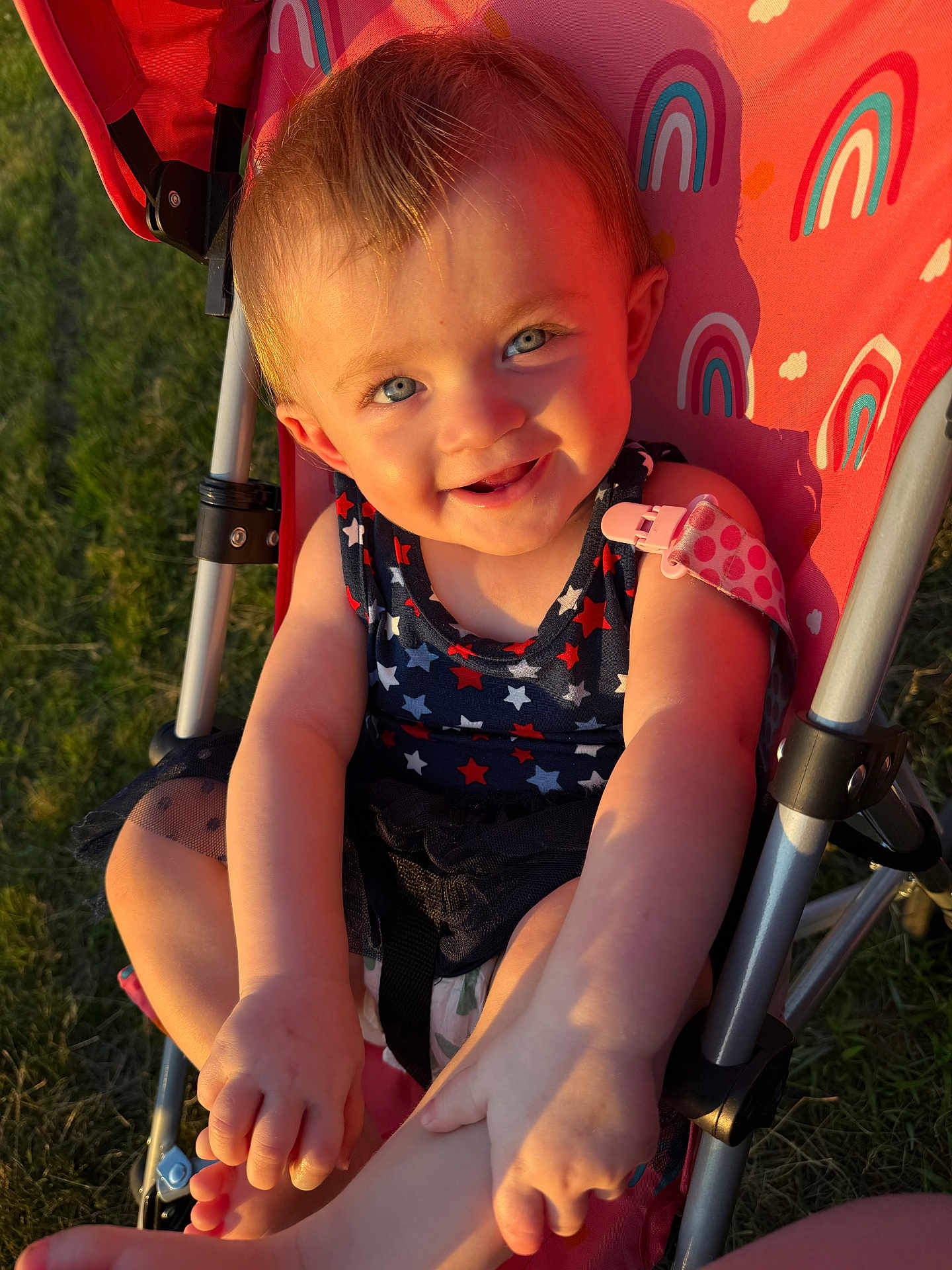 Oakley is registered to the contest to win money with this photo: toddler, child, stroller, outdoor, sunlight, grass, blue_eyes, smiling, happy, playing, summer, rainbow_pattern, clothing, skin, baby, foot, hand, seat, closeup, portrait