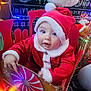 baby, child, santa_hat, red_clothing, holiday, christmas_lights, festive, toy, decorations, indoor, cheerful, cute, holiday_season, winter, celebration, sleigh, plush, smiling, face, seasonal
