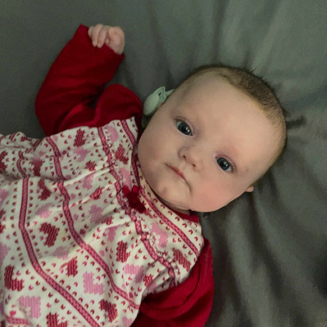 Lottie-May joined the competition — help win amazing prizes! baby, blanket, child, clothing, cute, expression, eyes, face, hand, head, indoor, infant, lying_down, patterned_dress, portrait, purple_toy, red_sleeves, skin, toy, young