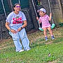 child, woman, swing, playground, grass, flowers, tree, fence, cap, glasses, pink_clothing, casual_wear, outdoor, smiling, happy, nature, daylight, fun, leisure, person