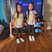 Harmony is registered to the contest to win money with this photo: child, twin, girl, smiling, standing, backpack, matching_outfits, television, living_room, wood_floor, indoor, black_pants, sneakers, long_hair, curtains, furniture, technology, happy, person, home