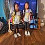 child, twin, girl, smiling, standing, backpack, matching_outfits, television, living_room, wood_floor, indoor, black_pants, sneakers, long_hair, curtains, furniture, technology, happy, person, home