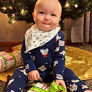 Luke is registered to the contest to win money with this photo: baby, infant, child, christmas_tree, ornament, string_lights, present, gift, wrapping_paper, green_ribbon, bib, onesie, smile, sitting, carpet, holiday, festive, indoor, tree_skirt, cute