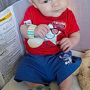 Koda is registered to the contest to win money with this photo: baby, child, playpen, red_shirt, blue_shorts, dinosaur_slippers, toy, indoor, home, floor, wood, curious, seated, comfort, infant, cute, casual_clothing, person, face, soft_lighting
