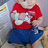 baby, child, playpen, red_shirt, blue_shorts, dinosaur_slippers, toy, indoor, home, floor, wood, curious, seated, comfort, infant, cute, casual_clothing, person, face, soft_lighting