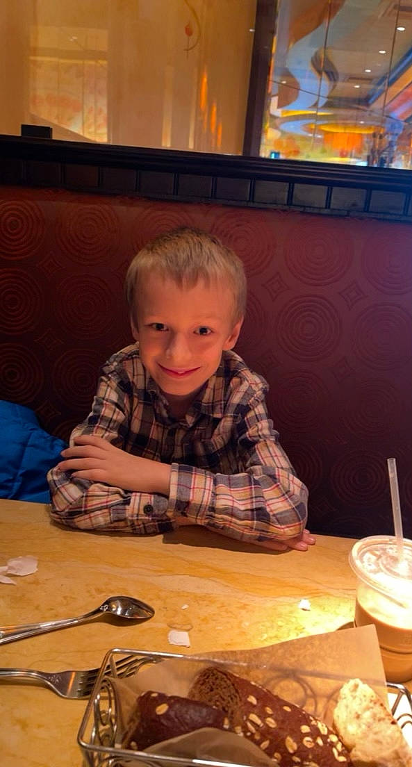 Tyson is registered to the contest to win money with this photo: audio_equipment, drink, event, flooring, fun, glass, hardwood, ingredient, joy, pattern, person, room, sitting, smile, t_shirt, table, tableware, toddler, toy, varnish