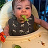 Hollis is registered to the contest to win money with this photo: baby, child, toddler, high_chair, broccoli, vegetables, eating, food, portrait, face, smiling, indoor, kitchen, bib, dinosaur_pattern, green_food, messy_meal, infant, seated, playful