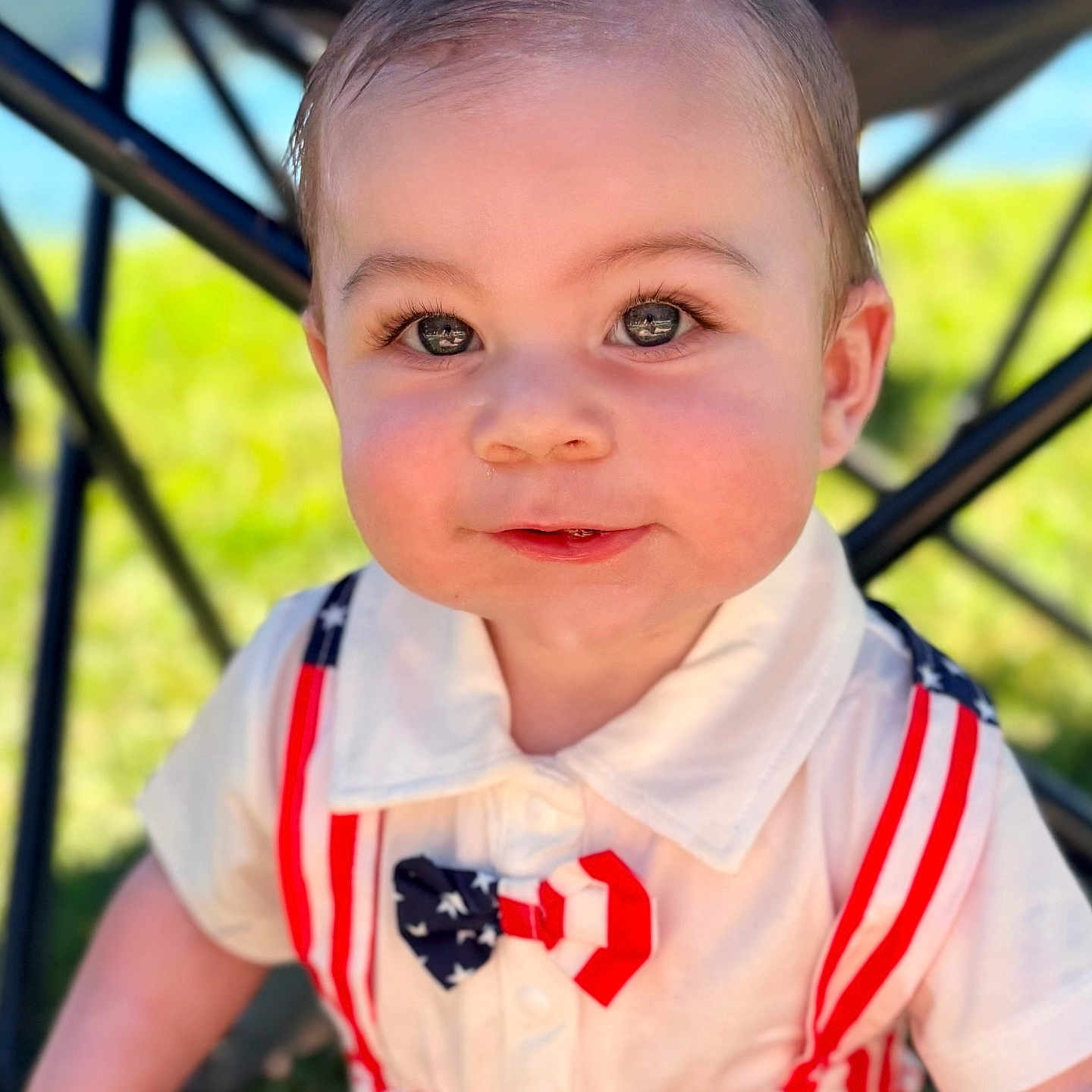 Hollis is registered to the contest to win money with this photo: baby, child, infant, portrait, smiling, face, eyes, outdoor, sunlight, patriotic_clothing, red_white_blue, dress, collar, stripes, seated, stroller, chair, grass, close_up, happy