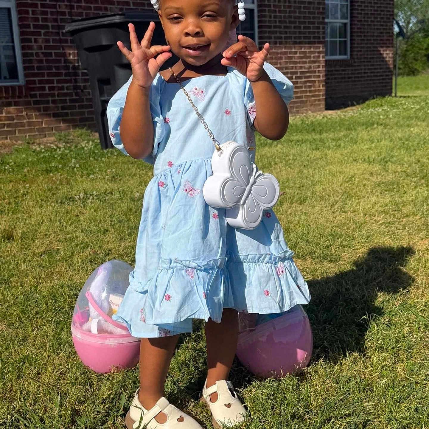 De' Azhia is registered to the contest to win money with this photo: child, toddler, girl, blue_dress, outdoor, grass, sunny, shoes, bag, butterfly, pose, playful, smile, hair_beads, brick_house, window, shadow, plastic_egg, toy, accessory