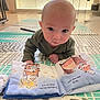 baby, infant, tummy_time, bald_baby, face, eyes, play_mat, picture_book, floor, indoor, crawling, curious, portrait, hand, cheek, onesie, living_room, bright_light, toy_book, close_up