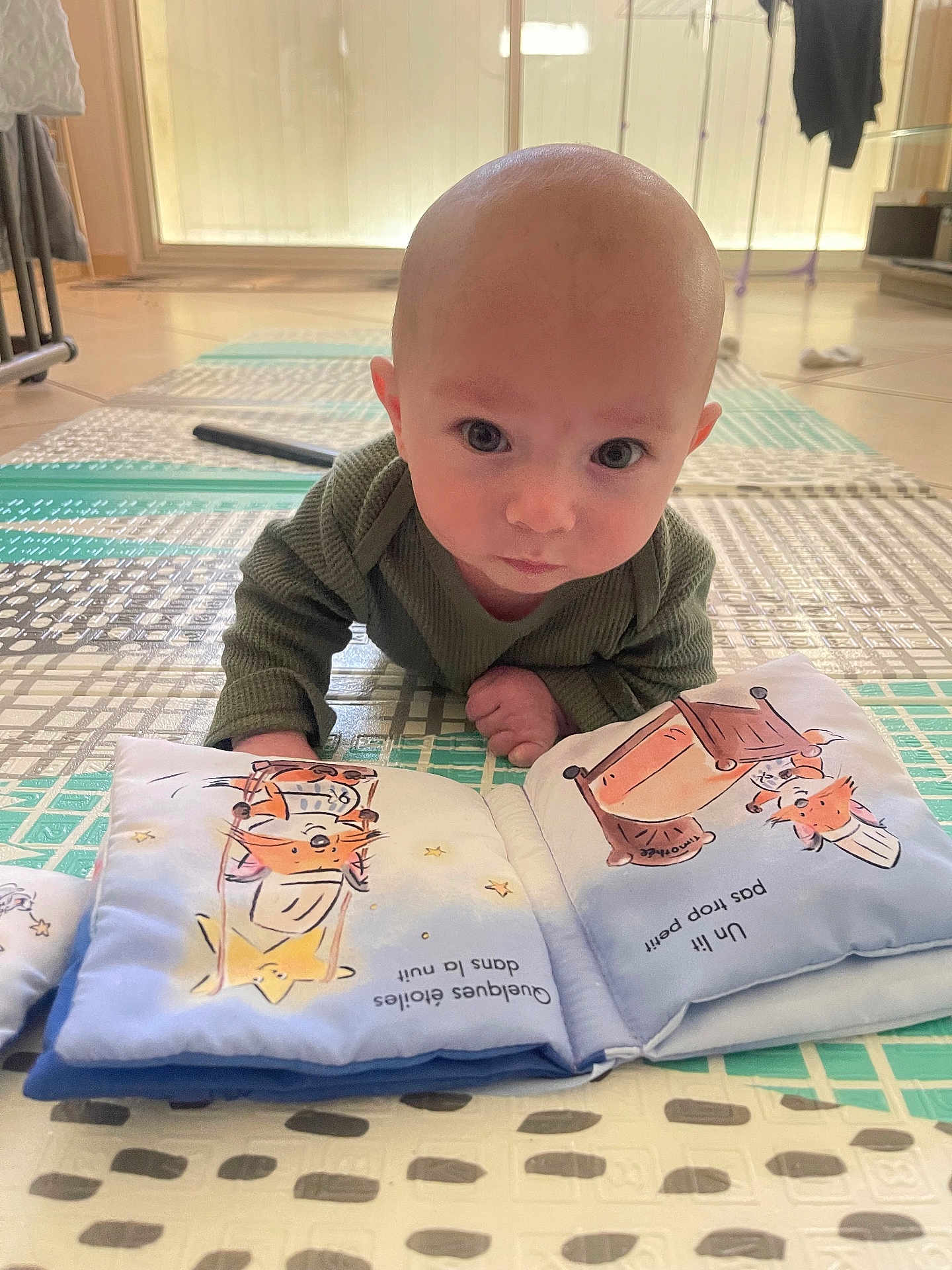 Matheo a rejoint le concours — aidez-le/la à gagner de superbes lots ! baby, infant, tummy_time, bald_baby, face, eyes, play_mat, picture_book, floor, indoor, crawling, curious, portrait, hand, cheek, onesie, living_room, bright_light, toy_book, close_up