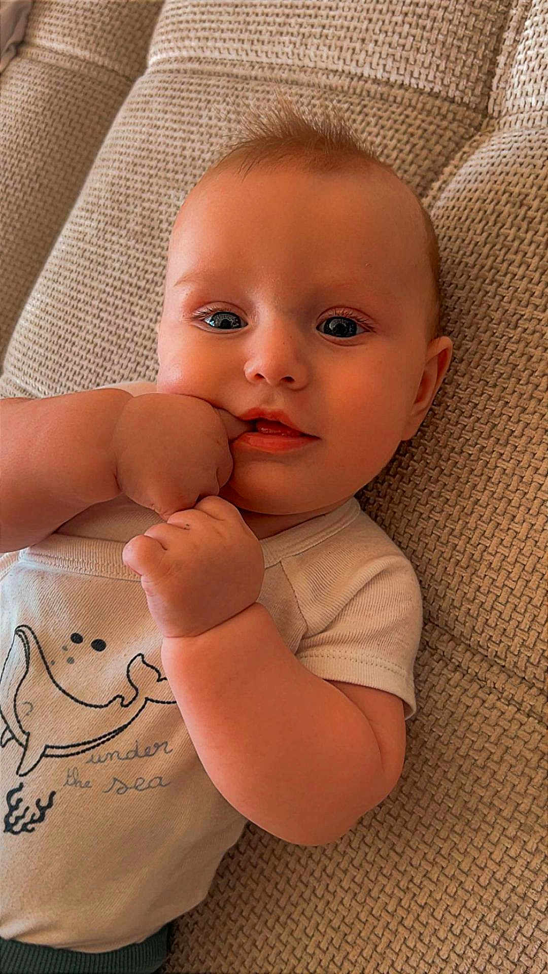 Lèven a rejoint le concours — aidez-le/la à gagner de superbes lots ! infant, baby, child, face, eyes, hand, fingers, cheek, mouth, onesie, clothing, couch, fabric_texture, portrait, close_up, lying_down, baby_smile, adorable, skin, indoor