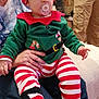 Lèven a rejoint le concours — aidez-le/la à gagner de superbes lots ! baby, child, pacifier, elf_hat, elf_costume, striped_leggings, red_and_white_stripes, green_top, sitting_on_lap, adult_hand, couch, fireplace, holiday_decorations, christmas, living_room, cozy, toddler, plush_boots, face, portrait