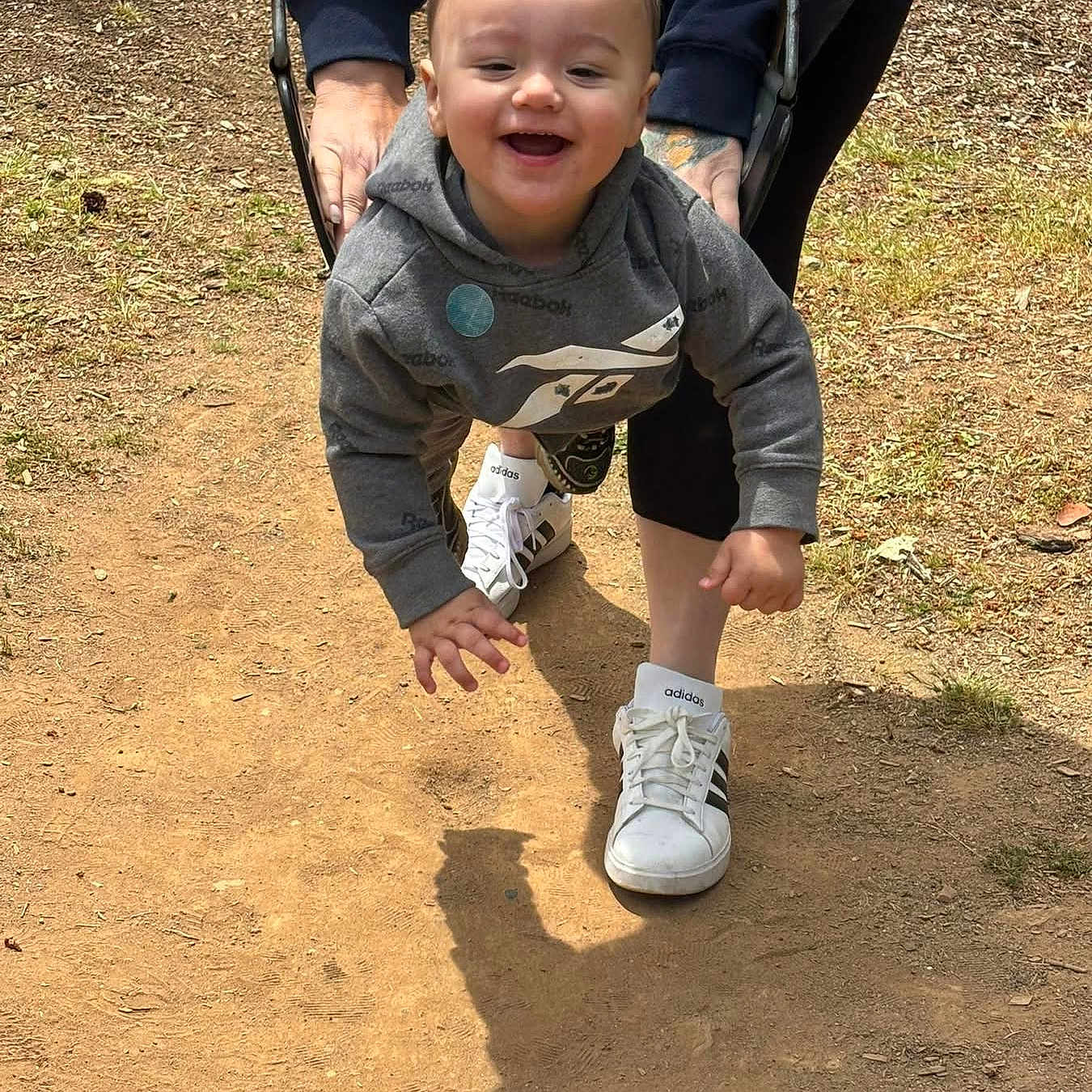 Jaden is registered to the contest to win money with this photo: child, toddler, swing, playground, outdoor, smile, happy, person, sneakers, adidas, hoodie, adult_hands, shadow, dirt_path, grass, sunlight, casual_clothing, fun, playing, nature
