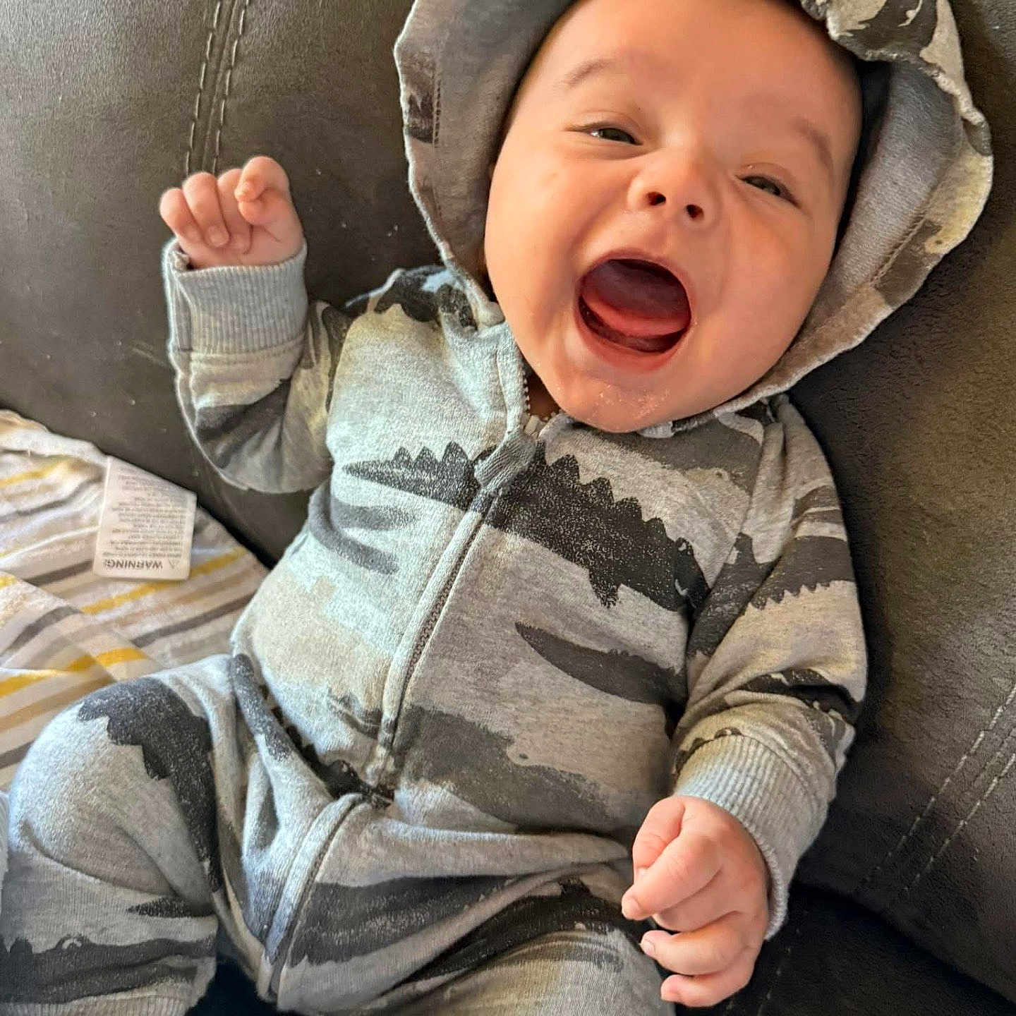 Jordin is registered to the contest to win money with this photo: baby, infant, smiling, onesie, hoodie, dinosaur_print, couch, leather, happy, child, indoor, cute, young, person, seated, clothing, playful, joy, fist, relaxed